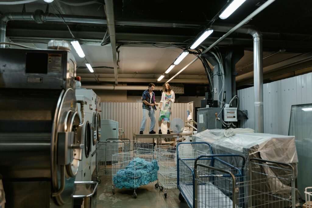 Two people working inside an industrial laundry facility with large machines and laundry carts.